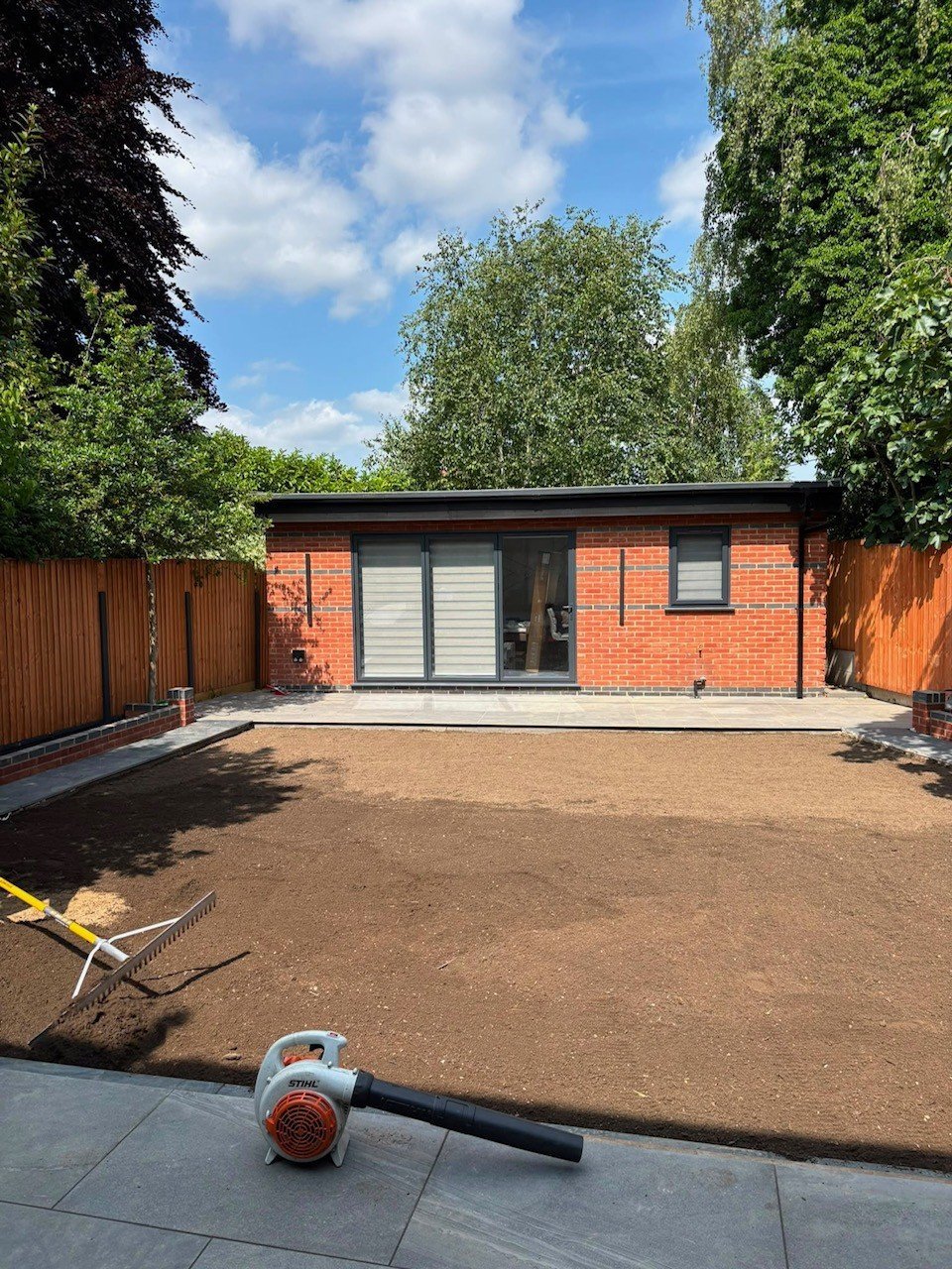 Modern brick garden room with large sliding glass doors, surrounded by wooden fences and green trees, under blue sky