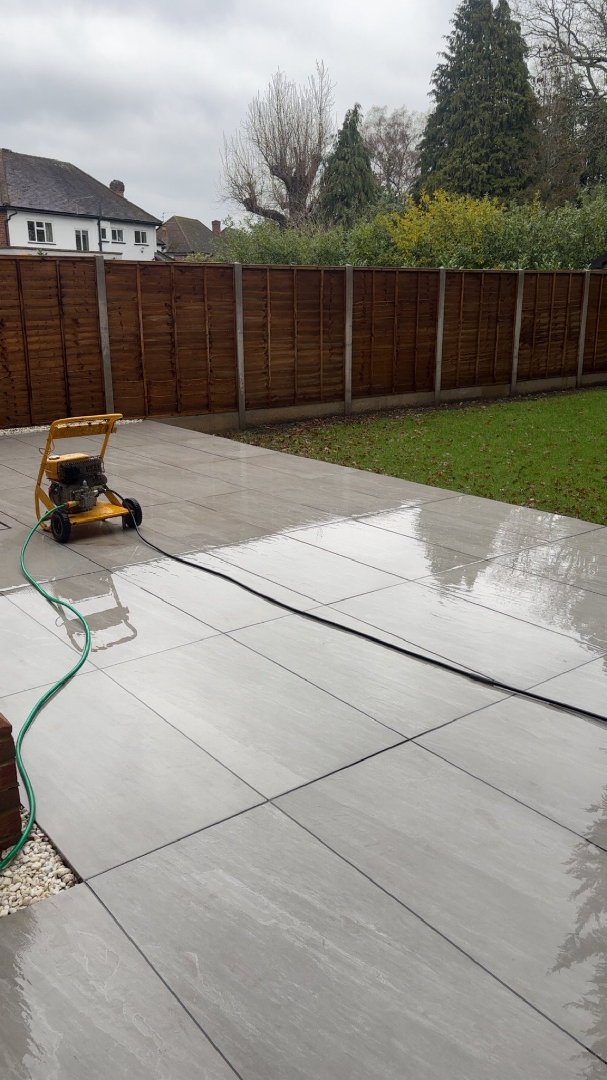 Wet patio being cleaned with pressure washer in a residential garden with brown fence and grass lawn