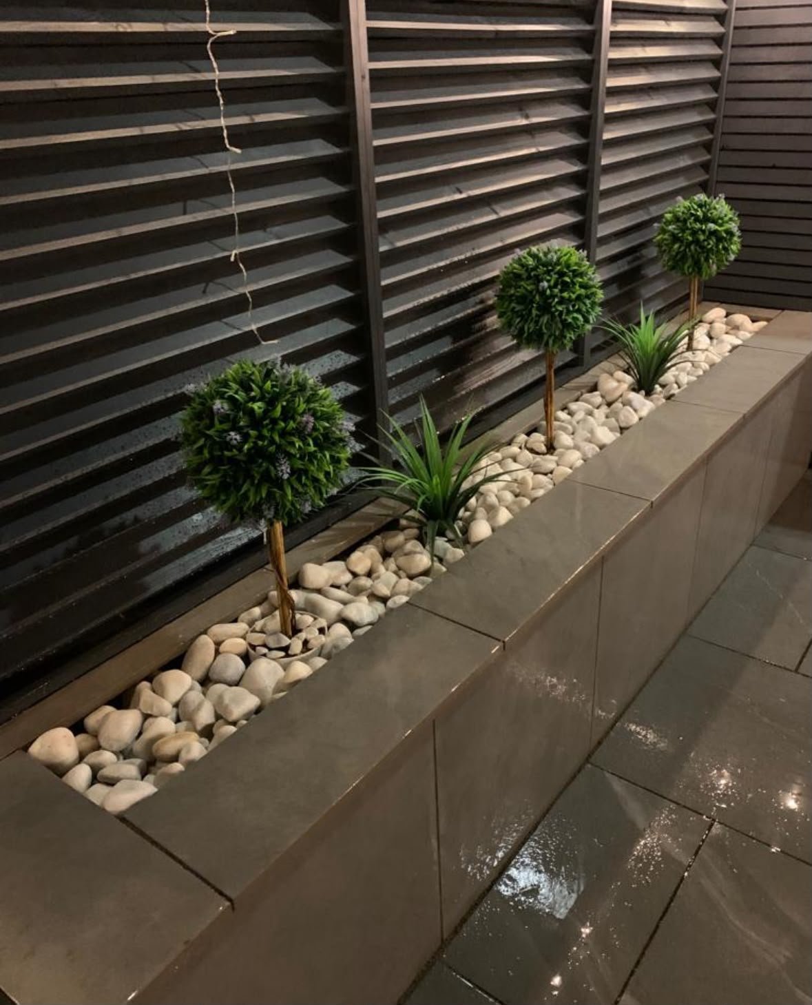 Modern garden planter with three potted topiaries and white river stones against horizontal black slatted fence
