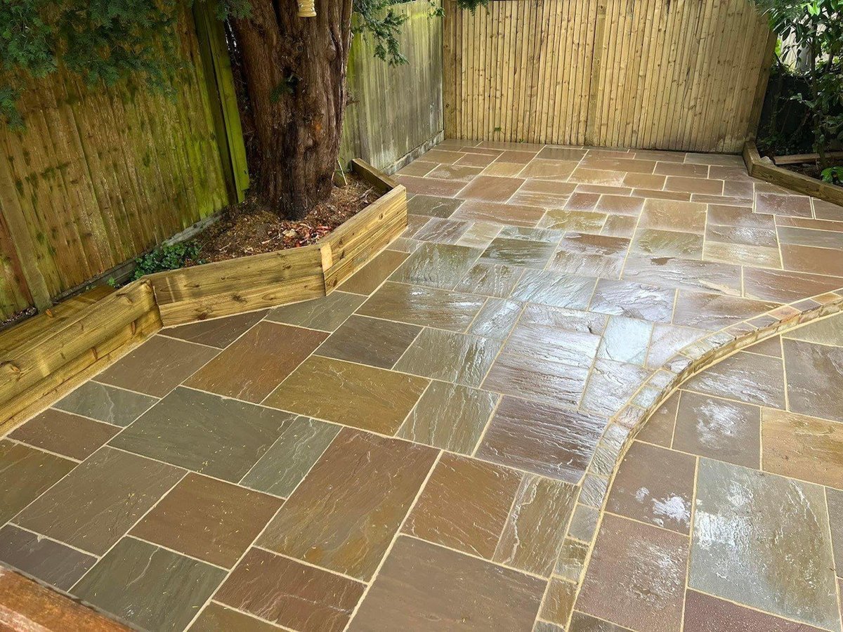 Newly installed stone patio with tan and brown mixed-color pavers in a herringbone pattern, bordered by wooden raised beds and fencing